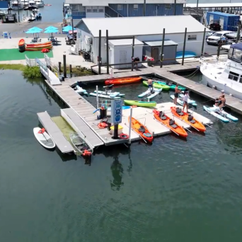 Marina with kayaks, paddleboards, and a dock beside a building and parking area.
