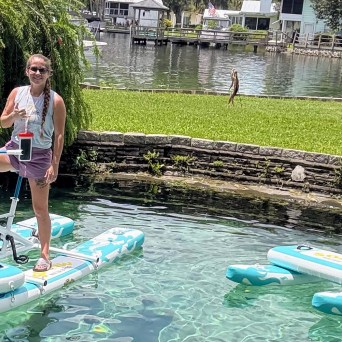 Two people riding water bikes on a clear water lake on a sunny day, with trees and houses in the background.